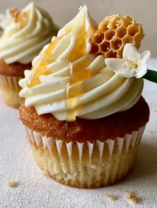 Eye-level shot of a single honey cupcake with golden-brown sponge and ivory honey buttercream on a linen surface. (honey cupcakes)