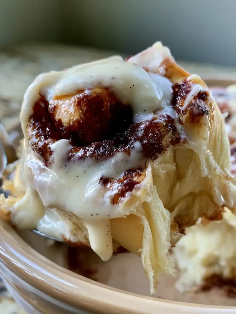 A 45-degree angle shot of cinnamon rolls in a ceramic dish with thick white frosting dripping into dark caramelized swirls.