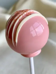 Macro eye-level shot of a single pale pink cake pop showing a high-gloss reflective sheen and a thick structural chocolate collar. (how to make cake pops)