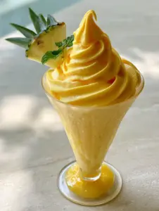 Close-up of a vibrant yellow pineapple soft serve with a matte finish and fresh mint garnish. (Dole Whip slush)