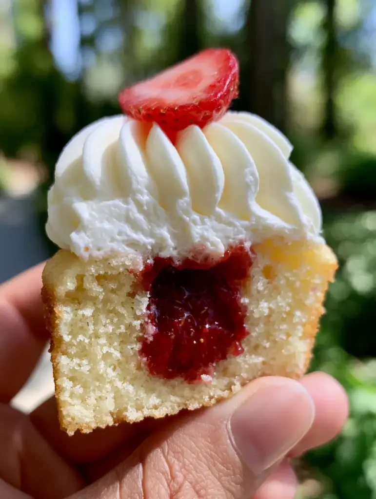 Strawberries and Cream Cupcakes With Ruby Red Strawberry Filling 2 Extreme close-up of a halved vanilla cupcake held in a hand, showing the deep ruby-red chunky strawberry compote center and airy whipped cream. (strawberries and cream cupcakes)