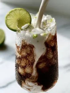 A POV shot of heavy coconut cream being poured into dark cola, creating white marble swirls in a glass with pebble ice. (dirty soda recipe)